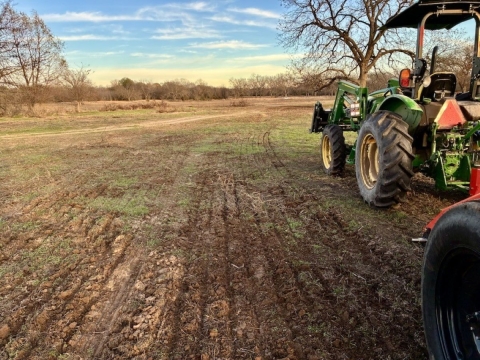 A field with freshly disturbed soil and green patches of new growth. A green tractor with large tires is parked on the right, and bare trees border the field beneath a blue sky with light clouds.