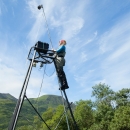 A person stands near the top of a tall ladder in a remote area surrounded by trees and greenery.