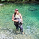 A service member wearing waders stands in a river holding a camera.