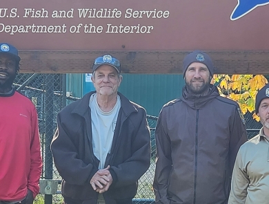 Four men stand smiling in front of a large sign for the Eagle Creek National Fish Hatchery. Sunlight and green grass are behind them.