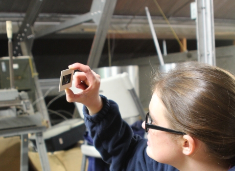A young woman in glasses holds a slide up to look at it in an attic storage space.