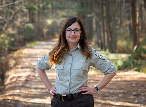 A woman with long brown hair stands on a trail framed by forest. Her hands are on her hips. She wears glasses and a U.S. Fish and Wildlife Service Uniform. 