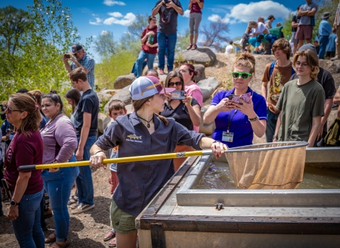 A high school student holding a net over a fish trailer. There are hundreds of people in the background as well as green trees and a blue sky with clouds.