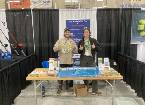 Two biologist stand smiling behind an outreach booth focused on invasive species prevention and identification in Alaska.