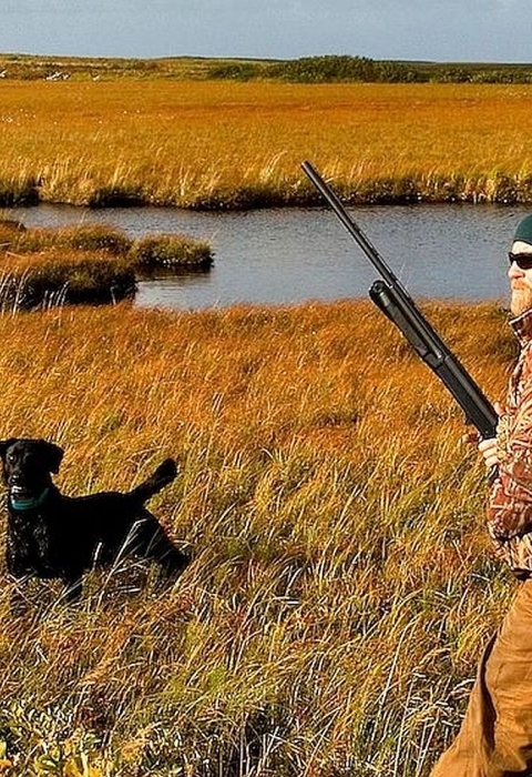 Two men carrying guns and backpacks walk across grassland next to a body of water with their hunting dog