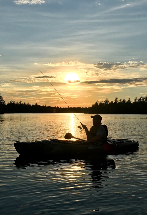 The silhouette of a person in a kayak holding a fishing rod while floating on a large body of water. The sun sets behind them. The sky is colored with vibrant blues and yellows.