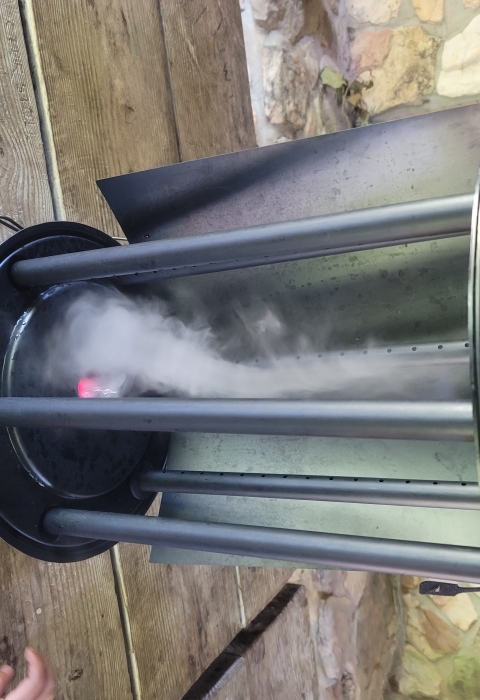 Two foot high hollow metal cylinder 10 inches in diameter sitting on top of a wood picnic table. There is tan mortared stonework behind the table and a youth hand next to the machine. The hollow cylinder is open in the front and smoke is swirling in an upside down vortex. 