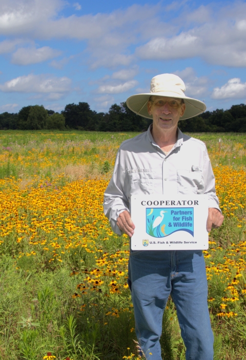 A person wearing a wide‑brimmed hat stands in a large field filled with yellow wildflowers. They hold a sign that reads “Cooperator – Partners for Fish & Wildlife – U.S. Fish & Wildlife Service.” Trees and a blue sky with scattered clouds appear in the background.