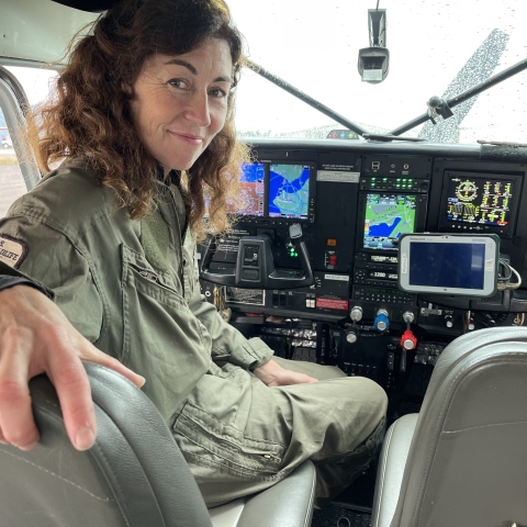 A women with brown hair looks back at the camera in the pilot seat of an airplane