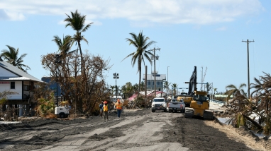 Heavy equipment parked on damaged asphalt alongside construction debris and damaged palm trees.