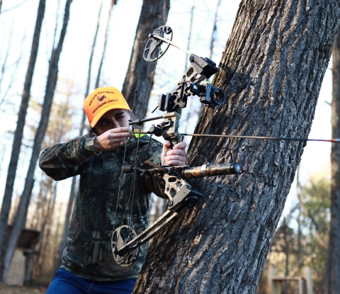 Lady hunting using a bow, wearing a camo jacket and orange hat. 