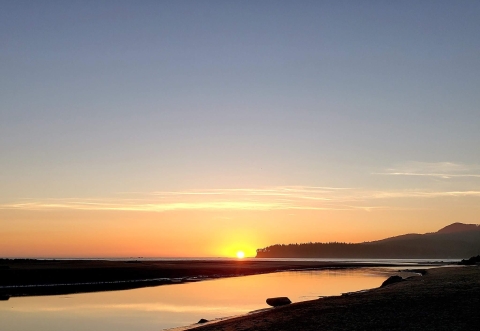 A view of a yellow and orange sky with the sun low on the horizon and a calm river mouth in the foreground.