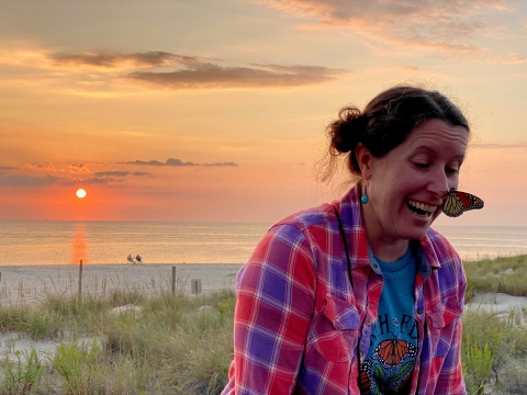 A person stands smiling on a sand dune as a butterfly sits on their nose