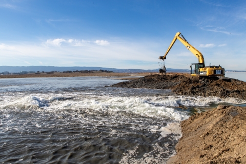 A bulldozer works to remove part of an earthen levee separating two bodies of water.