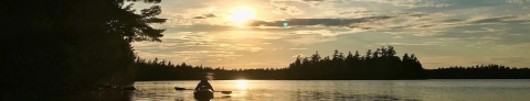 A kayaker paddling toward a setting sunset on a pond surrounded by evergreen trees