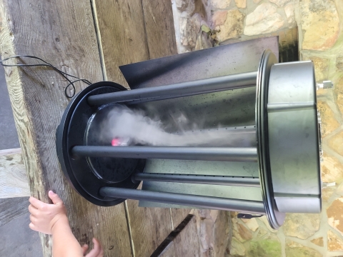 Two foot high hollow metal cylinder 10 inches in diameter sitting on top of a wood picnic table. There is tan mortared stonework behind the table and a youth hand next to the machine. The hollow cylinder is open in the front and smoke is swirling in an upside down vortex. 