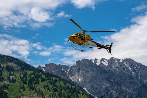 A yellow helicopter with a crewmember looking out soars above rocky mountains in Wyoming.