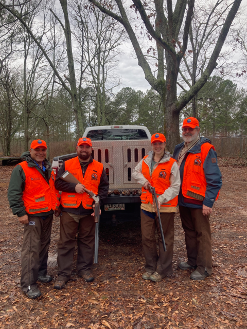 Four people in orange hunting vests, two with firearms in their hands, stand in front of a truck in the woods smiling