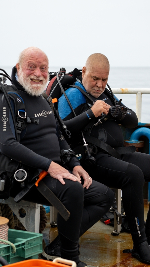 Two Caucasian men are doing a safety check of their dive equipment while sitting on the deck of the ship before they enter the water.