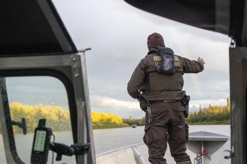 A Federal Wildlife Officer on the bow of a boat on a river waves at an oncoming boat. A rainbow is visible in the background. 