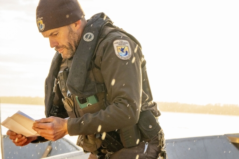A uniformed Federal Wildlife Officer checks a hunter's license and tag with some light rain and a sunny background. The shoreline of a river is visible and out of focus in the background. 