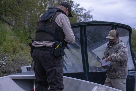 A uniformed Federal Wildlife Officer stands on his boat along side a hunter's boat where the hunter is standing holding paperwork. Both boats are on the shoreline of a river.