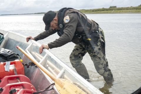 a uniformed officer pushes his boat off a shallow section while standing in the middle of a large river