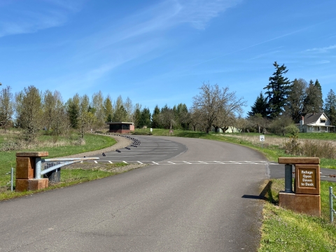 Image of an asphalt driveway with open metal entrance gates and a white striped crosswalk across the driveway and green grass on each side of the driveway.