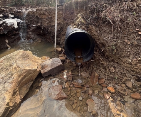 Image of metal pipe with water draining into muddy and rocky area.