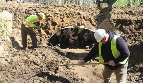 Two people in hard hats and vests shovel dirt in front of a metal culvert.