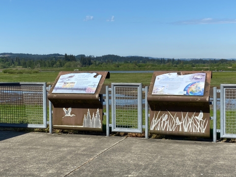 Image of two colorful signs attached to brown metal structures decorated with gray metal wildlife silhouettes and surrounded by a concrete pad, metal fence, hills, green plants and open water.