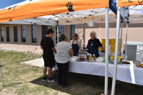 Two people stand behind a table, which contains hot dogs and chips, serving food to two adults