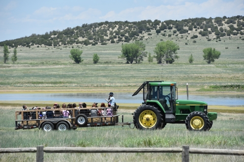 A tour-led tractor with a trailer carrying about a dozen people rides by a lake.