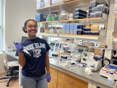 a young, black woman wearing glasses and a blue t-shirt that says Rhode Island holds a pipette while standing in front of a crowded laboratory counter