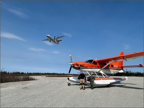 two people standing in front of a plane on the ground while another flies in the air behind them