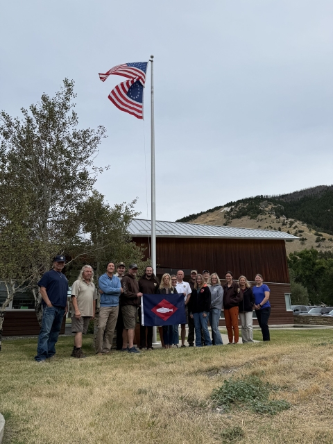 2 US flags fly and at base of flagpole, people pose with fish flag