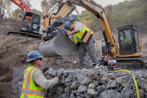 A person wearing a hard hat and a yellow safety vest stands beside a rock wall while another person similarly dressed unloads rocks from the bucket of an excavator