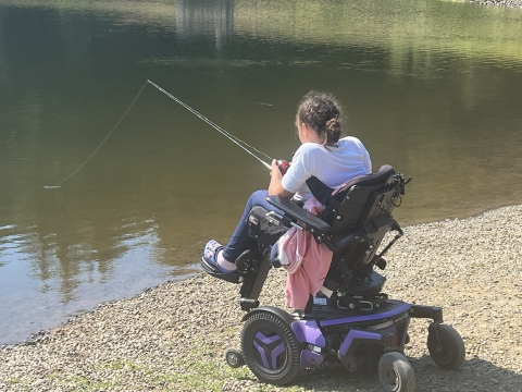 A child using a purple and black motorized wheelchair fishes along a pebbled shoreline beside still water.