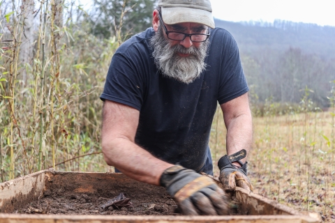 Man standing on the edge of a field, wearing work gloves, and sorting through dirt in a large sieve in front of him