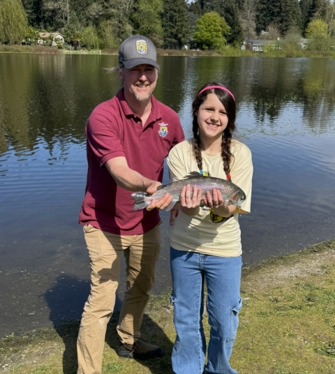 Service staff member and youth event participant posing with a rainbow trout. Lake, houses and trees in the background.