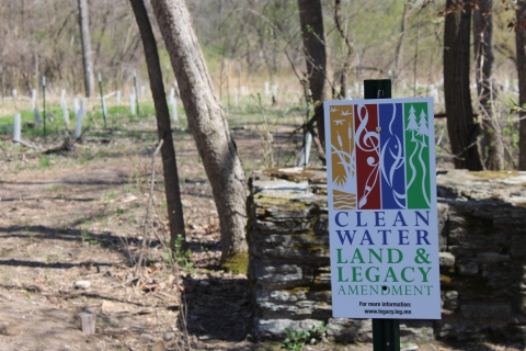A sign that reads “Clean water land & legacy amendment. For more information: www.legacy.leg.mn” There is a stone wall, trees and a dirt path behind the sign.