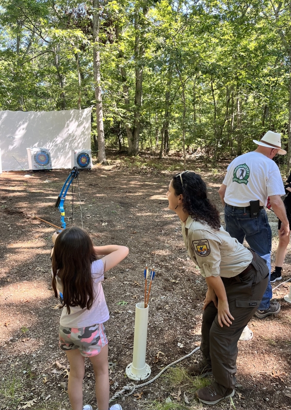 Ranger leans down as she's teaching a young girl how to shoot a bow and arrow at an archery range.