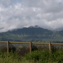 A tall fence cuts across a lush green landscape. There is a green mountain in the background with fluffy white clouds on it.