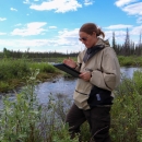 person with a clipboard looking at a patch of plants along a river