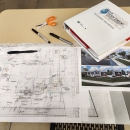 Image of a hand drawn blueprint, pictures of a house, binder, and pens on a des