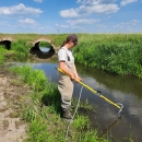 Woman in waders holds a pole and sticks it in the water. It is a summers day.