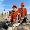 Two young brothers wearing orange and camo with the younger one holding a harvest mule deer by the antlers