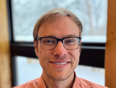 A man with light hair and glasses wearing an orange collared shirt smiles. Behind him is a window.
