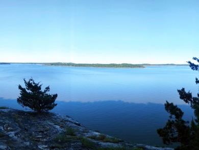 Panoramic view from lichen-covered bluff overlooking blue water and sky with green summer foliage in the distance