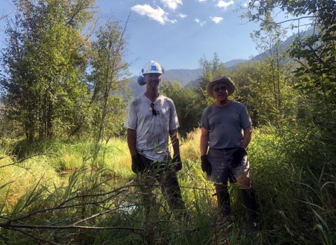 A tall man in hardhat, dirty t-shirt, and ear protection stands next to a shorter man in straw hat, shorts, and t-shirt, both wearing work gloves, in a field.